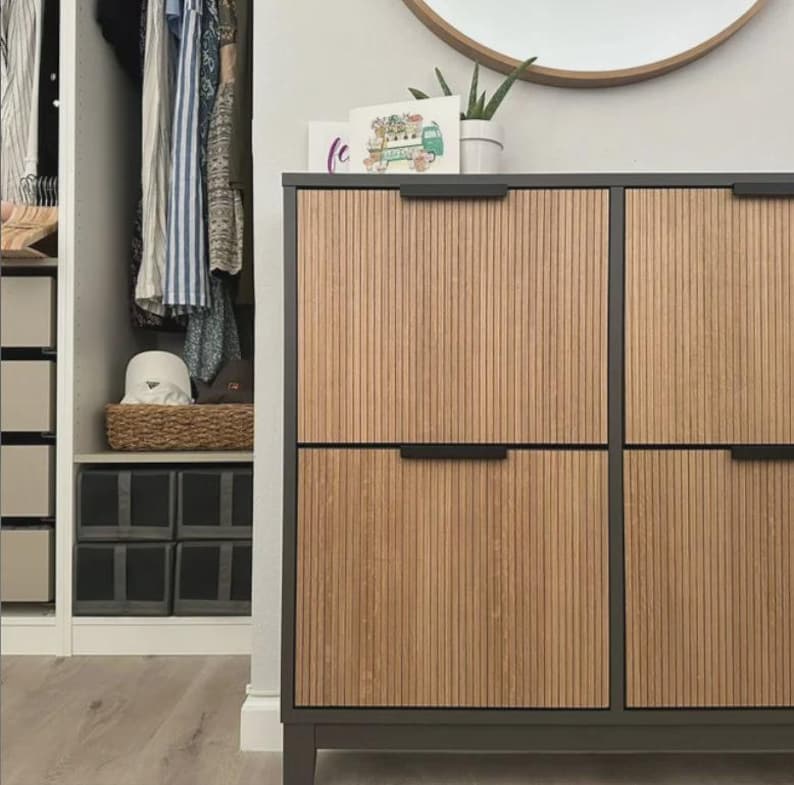 Op de afbeelding: Een rechthoekige opbergkast met een donkergrijs frame en vier lichthouten deuren met verticale latten en dunne zwarte horizontale handgrepen. De kast staat in een kamer met een kledingkast met hangende kleding en opbergbakken.
