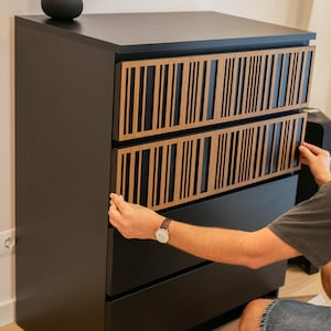 May include: A black dresser featuring four drawers, with two of the top drawer fronts covered by decorative light brown wood panels. These panels display a vertical slat pattern with varying widths, creating a textured, linear aesthetic.