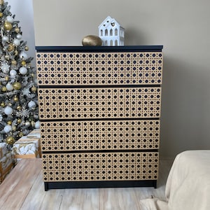 May include: A black four-drawer dresser featuring light tan woven cane panels on the front of each drawer. The dresser has a rectangular frame and a flat top surface. A small white house-shaped ornament and a gold sphere sit on the top.