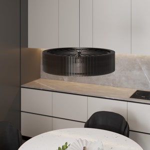 Pu&ograve; includere: Una lampada a sospensione circolare nera caratterizzata da un design a tamburo con lamelle verticali che si irradiano da un punto centrale. La lampada pende da un cavo sottile, mostrando una struttura aperta con una lampadina visibile all'interno.