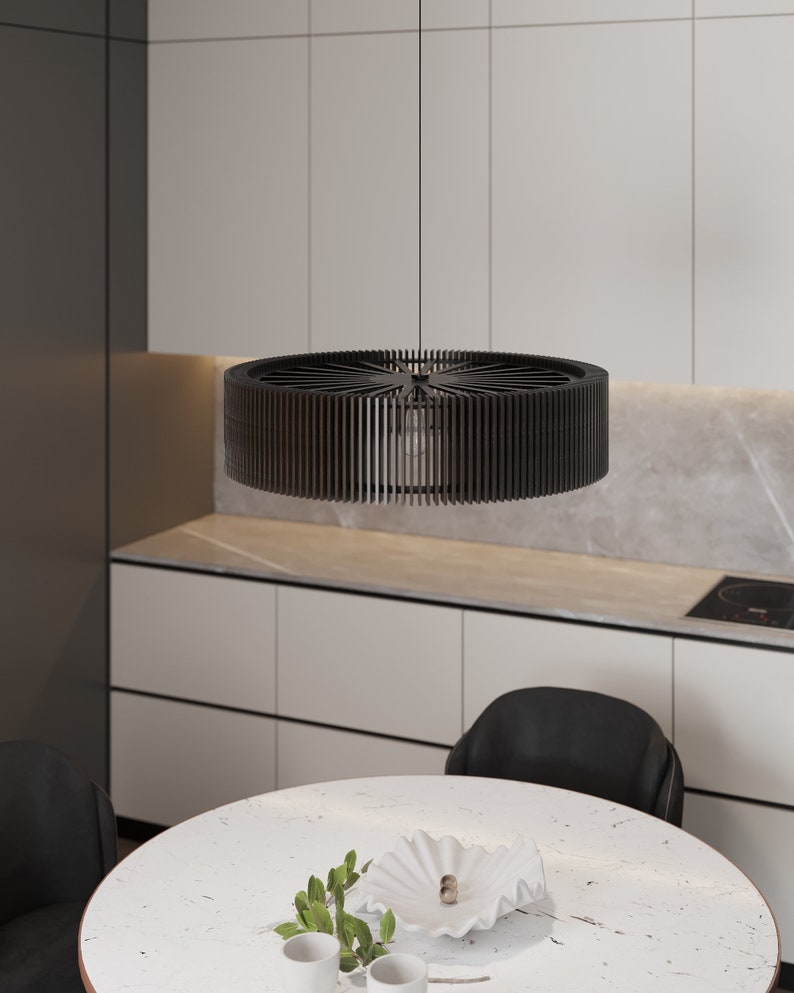 Pu&ograve; includere: Una lampada a sospensione circolare nera caratterizzata da un design a tamburo con lamelle verticali che si irradiano da un punto centrale. La lampada pende da un cavo sottile, mostrando una struttura aperta con una lampadina visibile all'interno.