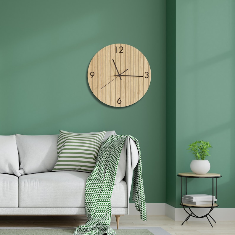 May include: A circular wall clock with a light wood grain finish featuring vertical slats and black numerals at the 12, 3, 6, and 9 o'clock positions. The clock has three thin black hands and is mounted on a solid green wall above a white sofa.