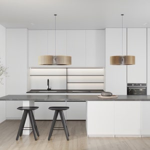 Pu&ograve; includere: Un interno cucina con armadi bianchi, un piano di lavoro grigio scuro e due sgabelli neri. Due lampade a sospensione cilindriche in legno pendono sopra una lunga isola. Un rubinetto nero &egrave; montato sul lavello e si nota un forno a incasso.