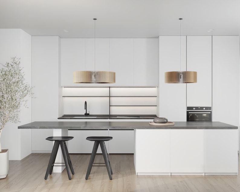 Pu&ograve; includere: Un interno cucina con armadi bianchi, un piano di lavoro grigio scuro e due sgabelli neri. Due lampade a sospensione cilindriche in legno pendono sopra una lunga isola. Un rubinetto nero &egrave; montato sul lavello e si nota un forno a incasso.