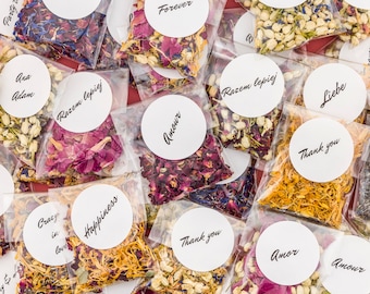 May include: Assortment of small clear bags filled with dried flower petals. Each bag has a white circle sticker with a different word or phrase written in black, such as "Forever", "Glück", "Amour", "Thank you", and "Razem lepiej".
