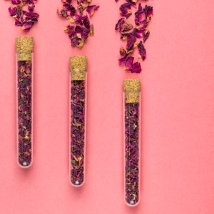 May include: Three glass test tubes with cork tops filled with dried rose petals on a pink background.