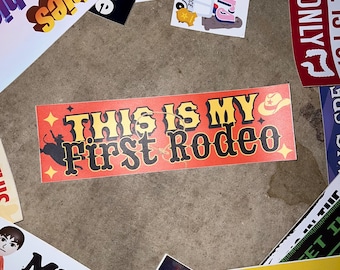 May include: A rectangular sticker with an orange-to-red gradient background featuring the text "THIS IS MY First Rodeo" in black and white lettering, accompanied by a small cowboy hat icon and a silhouette of a bull rider.