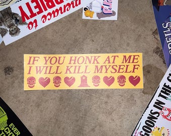 May include: A rectangular yellow bumper sticker featuring the red text IF YOU HONK AT ME I WILL KILL MYSELF, followed by a row of alternating red skull, heart, and tombstone icons printed across the bottom of the sticker.