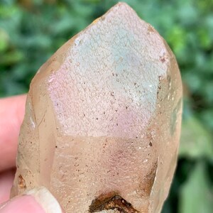 Puede incluir: Cristal de cuarzo marrón claro con una superficie texturizada y una punta afilada. El cristal tiene un aspecto natural y en bruto, con sutiles variaciones de color y una pequeña inclusión oscura cerca de la base. El fondo es verde borroso.