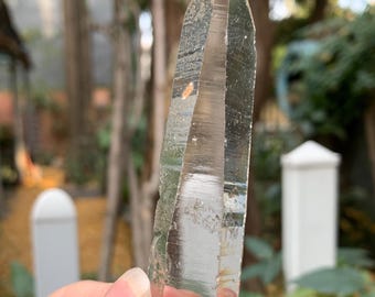 May include: A clear quartz crystal point with a light brown hue. The crystal is held in a hand and is in focus against a blurred outdoor background. The crystal has a sharp, pointed tip and visible internal structures.