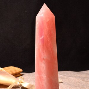 May include: A pink rose quartz crystal point, also known as a wand, is a natural crystal with a smooth, polished surface. The crystal is approximately 10 inches tall and has a pointed tip.