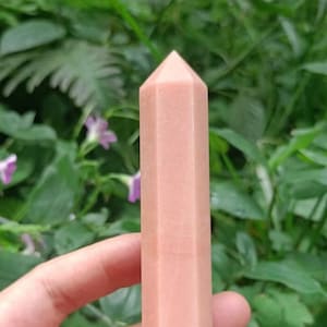 May include: A light pink, faceted crystal point, also known as a crystal wand, is held in a hand against a green leafy background.