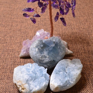 Puede incluir: Un árbol de amatista envuelto en alambre con un tronco de cobre se asienta sobre una base de tres cristales de celestita azules. Los cristales tienen forma de corazón y una textura rugosa y natural.