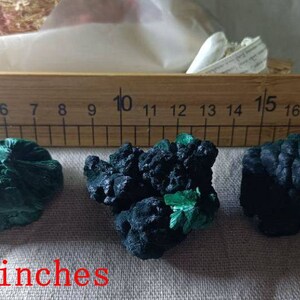 May include: Three green and black malachite mineral specimens, measuring between 1.4 and 2 inches in size, are displayed on a fabric surface next to a wooden ruler.