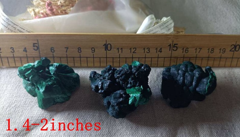 May include: Three green and black malachite mineral specimens, measuring between 1.4 and 2 inches in size, are displayed on a fabric surface next to a wooden ruler.