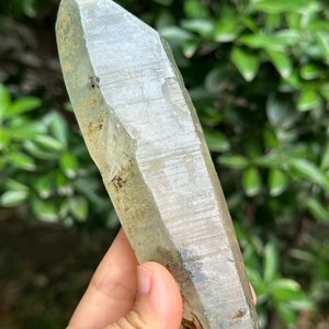 May include: A large, clear quartz crystal with a pointed tip and a rough, natural surface. The crystal has a slight green tint and is partially covered in a layer of brown and grey inclusions.