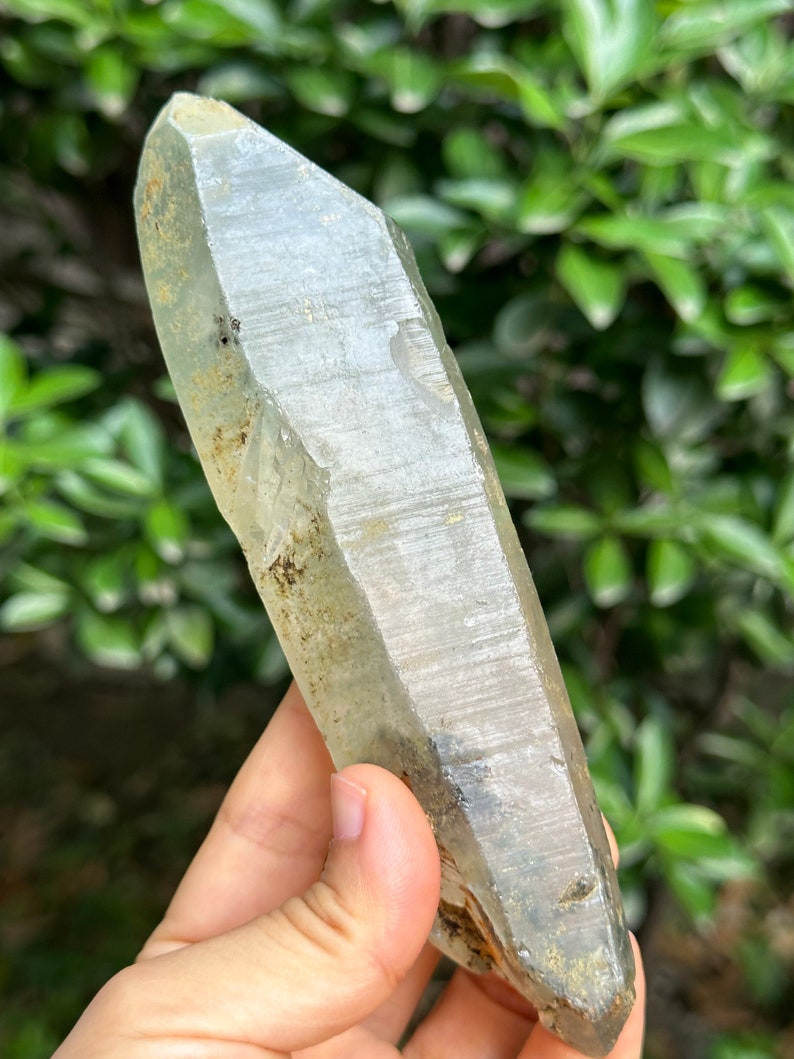 May include: A large, clear quartz crystal with a pointed tip and a rough, natural surface. The crystal has a slight green tint and is partially covered in a layer of brown and grey inclusions.