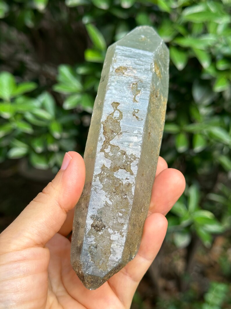May include: A clear quartz crystal point with a light grey, cloudy appearance. The crystal has a natural, rough texture and is approximately 15 cm long.