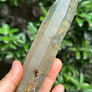 May include: A large, clear quartz crystal with a green hue and a rough, natural texture. The crystal is pointed at both ends and has a cloudy, milky appearance.