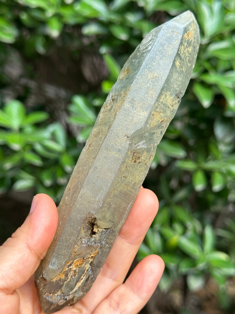 May include: A large, clear quartz crystal with a green hue and a rough, natural texture. The crystal is pointed at both ends and has a cloudy, milky appearance.