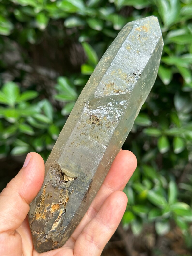 May include: A large, clear quartz crystal with a green hue and brown inclusions. The crystal is pointed at both ends and has a rough, natural texture.