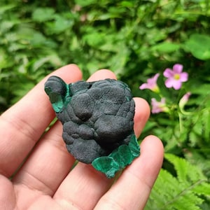 May include: A dark green malachite stone with a rough, textured surface. The stone has a smooth, polished edge.
