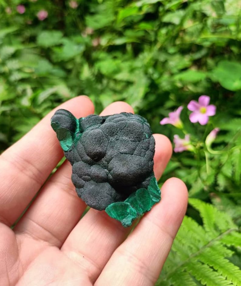 May include: A dark green malachite stone with a rough, textured surface. The stone has a smooth, polished edge.