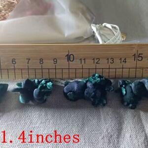 May include: Five green and black malachite stones, ranging in size from 0.8 to 1.4 inches, are displayed on a white fabric surface next to a wooden ruler.