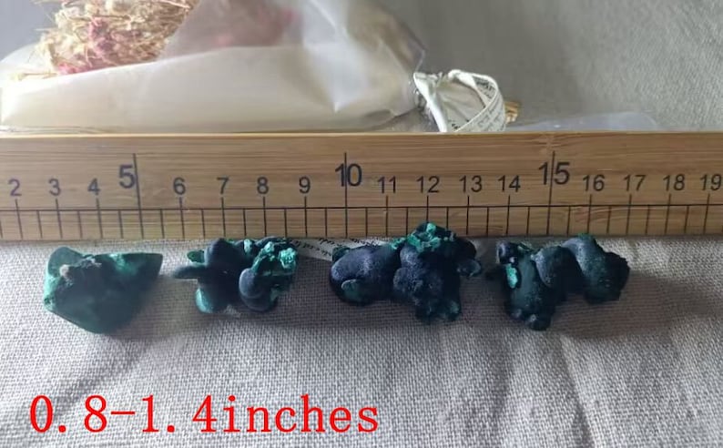 May include: Five green and black malachite stones, ranging in size from 0.8 to 1.4 inches, are displayed on a white fabric surface next to a wooden ruler.