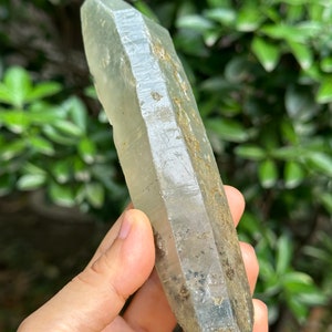 May include: A large, clear quartz crystal with a pointed tip and a rough, natural surface. The crystal has a light green hue and is partially covered in brown and black inclusions.