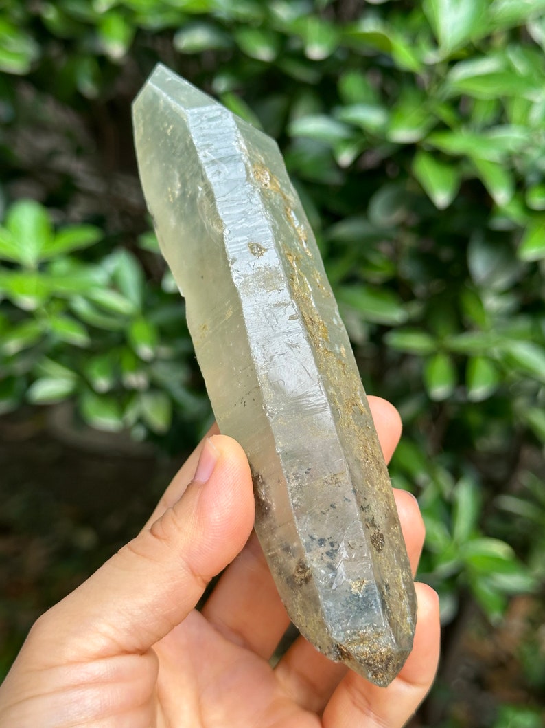 May include: A large, clear quartz crystal with a pointed tip and a rough, natural surface. The crystal has a light green hue and is partially covered in brown and black inclusions.