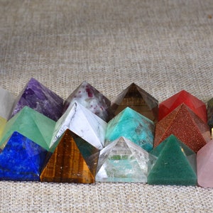 May include: A collection of 15 small crystal pyramids in various colors, including red, yellow, green, blue, purple, and white. The pyramids are arranged in a semi-circle on a textured surface.