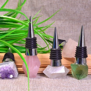 Può includere: Quattro tappi per bottiglie di vino con pietre preziose. Ogni tappo ha una parte superiore conica argentata e una base in gomma nera. Le pietre preziose sono ametista, quarzo rosa, quarzo trasparente e fluorite verde. Erba verde e bambù sullo sfondo.