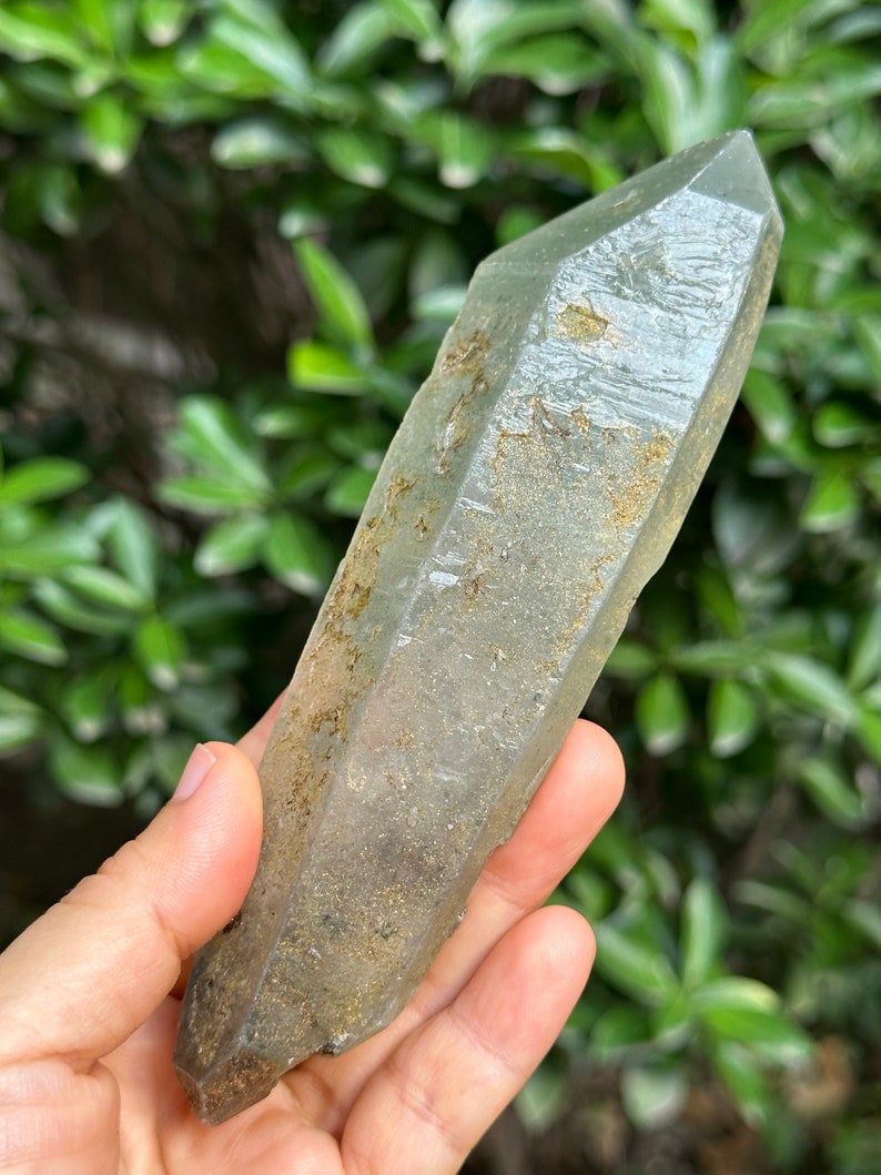 May include: A large, clear quartz crystal with a green hue and brown inclusions. The crystal is pointed at both ends and has a rough, natural texture.