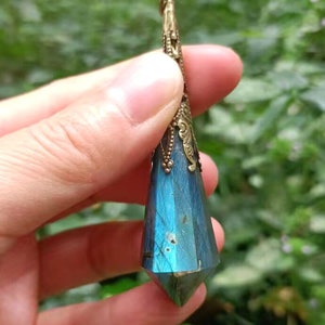 Op de afbeelding: Een blauwe en groene labradorit kristallen hanger met een goudkleurige metalen setting. De hanger is traanvormig en heeft een puntige punt.