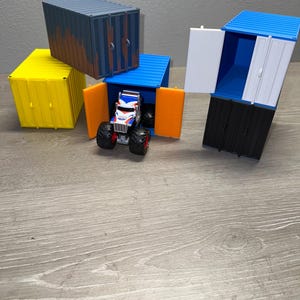 Könnte beinhalten: Ein Spielzeug-Monstertruck mit einem rot-weiß-blauen Design parkt in einem blauen Container mit orangen Türen. Weitere Container sind gelb und blau mit offenen Türen. Die Container stehen auf einer Holzoberfläche.