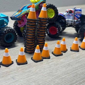 Può includere: Una collezione di monster truck giocattolo e coni stradali arancioni in miniatura con dettagli bianchi e neri. I camion sono dai colori vivaci con design dettagliati e ruote grandi. I coni sono disposti in pile e file.