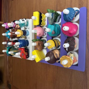 May include: A wall-mounted display rack, painted white and purple, holding a collection of colourful character figurines. The figurines are various shapes and sizes, with a variety of colours, including blue, yellow, pink, and brown. The rack is mounted on a wooden surface.