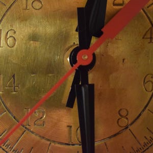 May include: Close-up of a brass clock face with black hands and a red second hand. The clock face has Roman numerals and minute markers. The hands point to approximately 10:07.