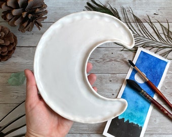 May include: A white ceramic crescent moon-shaped paint palette. The palette has a scalloped edge and is held in a hand. A watercolor painting of a night sky with a moon and two paint brushes are next to the palette.