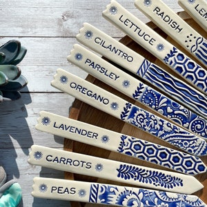 May include: Set of ten ceramic plant markers with blue and white designs. The markers are labeled with the names of herbs and vegetables, including: Lemon Balm, Radish, Lettuce, Cilantro, Parsley, Oregano, Lavender, Carrots, Peas.