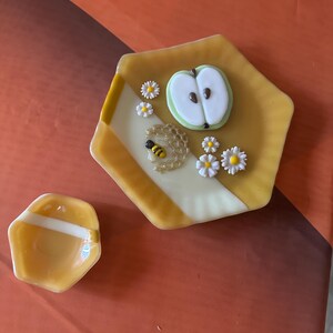 May include: Two decorative glass dishes. One is a small, hexagonal dish with a white stripe and a larger hexagonal dish with a yellow and white striped design. The larger dish has a bee, daisies, and an apple slice design.