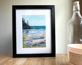 May include: A framed art print featuring a coastal landscape painting with a sandy beach, a small stream, driftwood, and evergreen trees on a cliffside. The scene includes small figures on the beach and in the water under a light blue sky, all enclosed in a simple black frame.