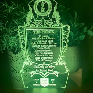 May include: Green acrylic light-up sign with the Army National Guard logo and the words "THE FORGE" and "SPC Emily McCaffery Fort Leonard Wood". The sign also includes a list of training exercises, including a 45-mile ruck march, 40 lbs ruck sack, night infiltration course, hand-to-hand combat, battle drills, land navigation, live fire exercise, obstacle course, sleep deprivation, and rite of passage.