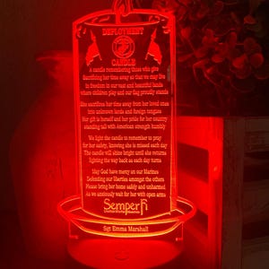 May include: A red illuminated candle with a white inscription. The inscription reads "Deployment Candle" and includes a quote about remembering those who serve in the military. The inscription also includes the text "Semper Fi" and "United States Marines".