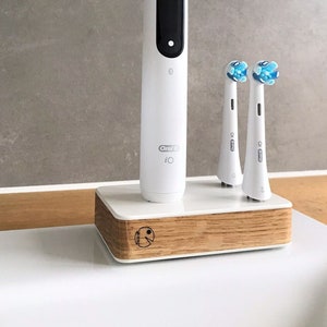 Könnte beinhalten: Eine weiße elektrische Zahnbürste mit einer Ladestation und zwei zusätzlichen Bürstenköpfen. Die Zahnbürste hat ein schwarzes Display und den Text "Oral-B iO" an der Seite. Die Ladestation ist weiß mit einem Holzsockel.