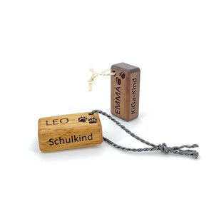 Può includere: Due etichette in legno con impronte di zampe e i nomi "Emma" e "Leo" incisi su di esse. L'etichetta di Emma dice "Kiga-Kind" e l'etichetta di Leo dice "Schulkind".