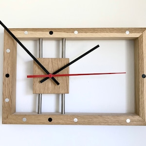 Könnte beinhalten: Eine Holzuhr mit quadratischem Zifferblatt und schwarzen und roten Zeigern. Die Uhr ist in einem rechteckigen Holzrahmen mit silbernen Akzenten und schwarzen Punkten gerahmt.