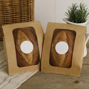 May include: Two brown paper bags with clear windows showcasing freshly baked loaves of bread. Each loaf has a white circular sticker. The bread has a golden-brown crust and a soft interior. The bags are on a wooden surface.