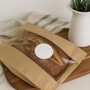 May include: A loaf of artisan bread in a brown paper bag with a clear window. A white circular sticker is on the bag. The bread sits on a wooden cutting board next to a white vase with green sprigs.